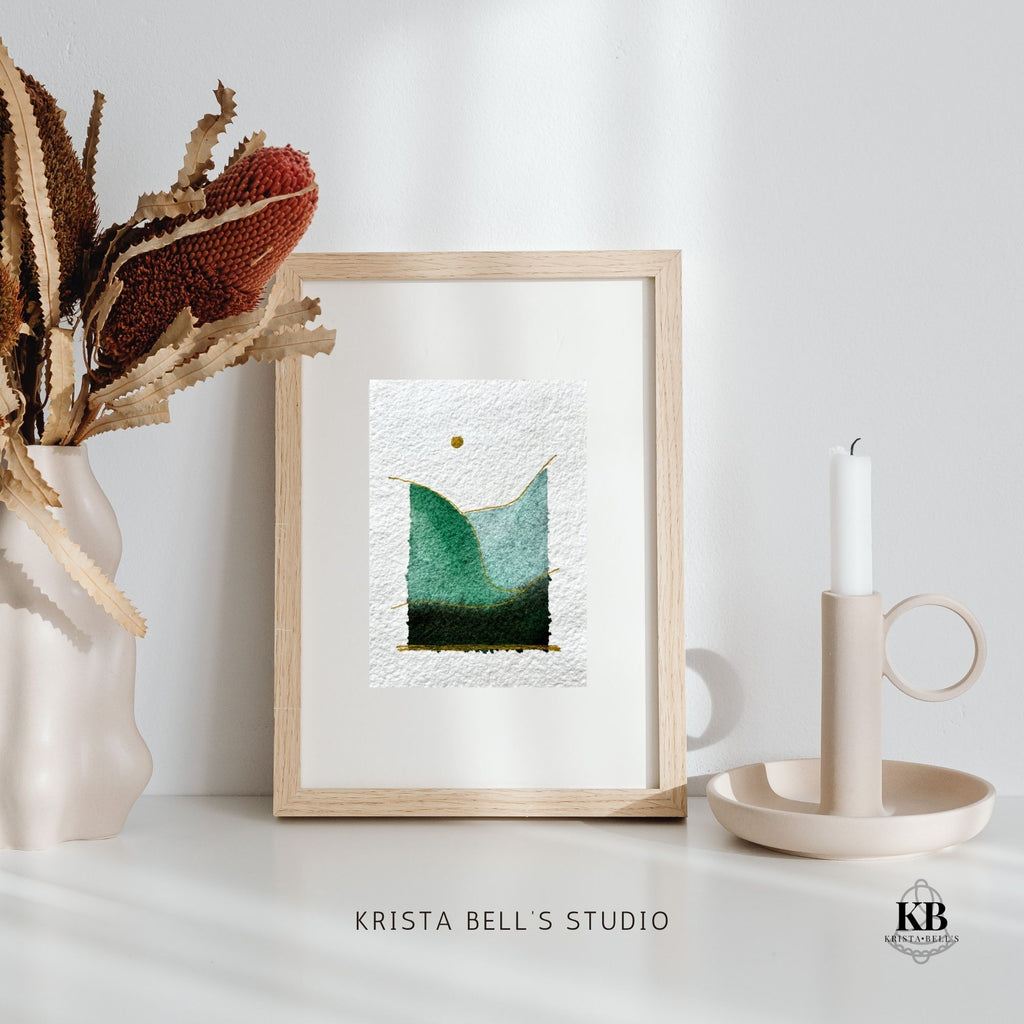Framed watercolor landscape artwork on a white surface with decorative elements, featuring 'Krista Bell's Studio' branding.