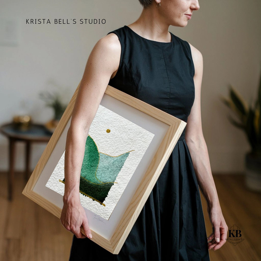Person holding a framed piece of art with a green abstract design, in a home setting.