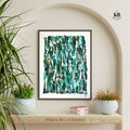 Framed abstract art print with green and blue patterns on a wall with plants and books below.