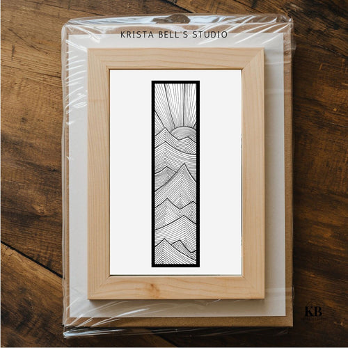 Framed artwork with a mountain design on a wooden surface, branded 'Krista Bell's Studio'.