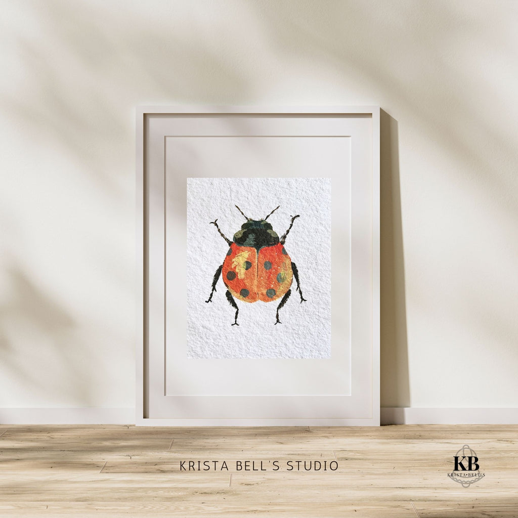 Framed artwork of a ladybug by Krista Bell's Studio on a wooden surface with a neutral background. A ladybug with reddish orange wings featuring black spots and a yellow highlight on a textured white background.