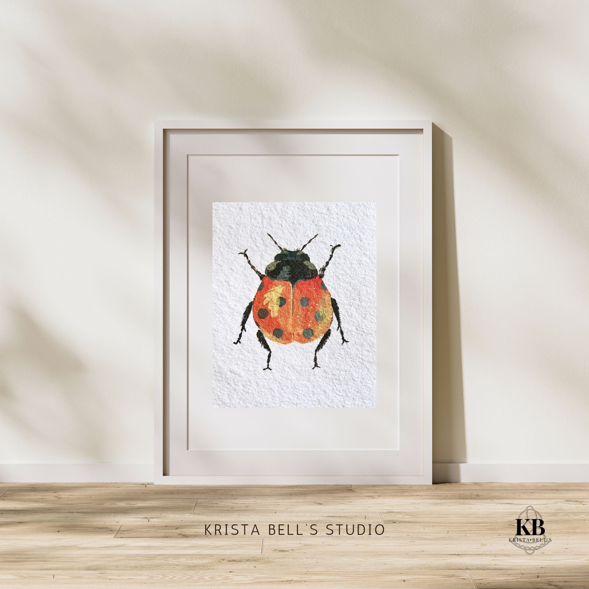Framed artwork of a ladybug by Krista Bell's Studio on a wooden surface with a neutral background. A ladybug with reddish orange wings featuring black spots and a yellow highlight on a textured white background.