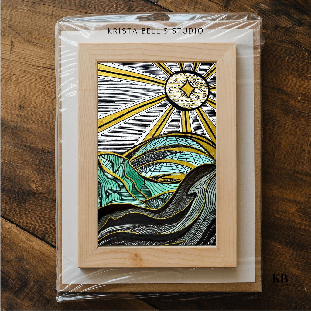Artistic depiction of a sun and mountains in a wooden frame, branded 'Krista Bell's Studio', on a wooden surface.