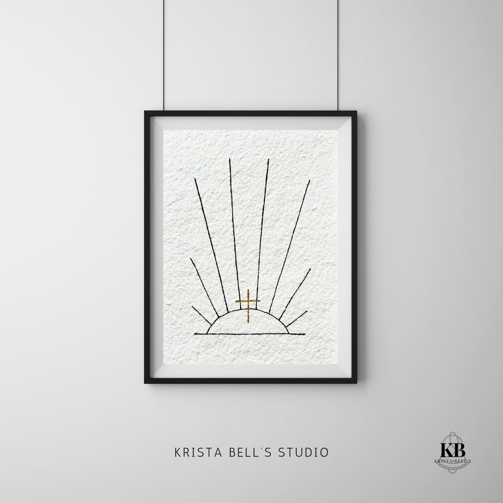 Framed artwork of black line sunrays and a golden cross on a gray wall, featuring 'Krista Bell's Studio' branding.