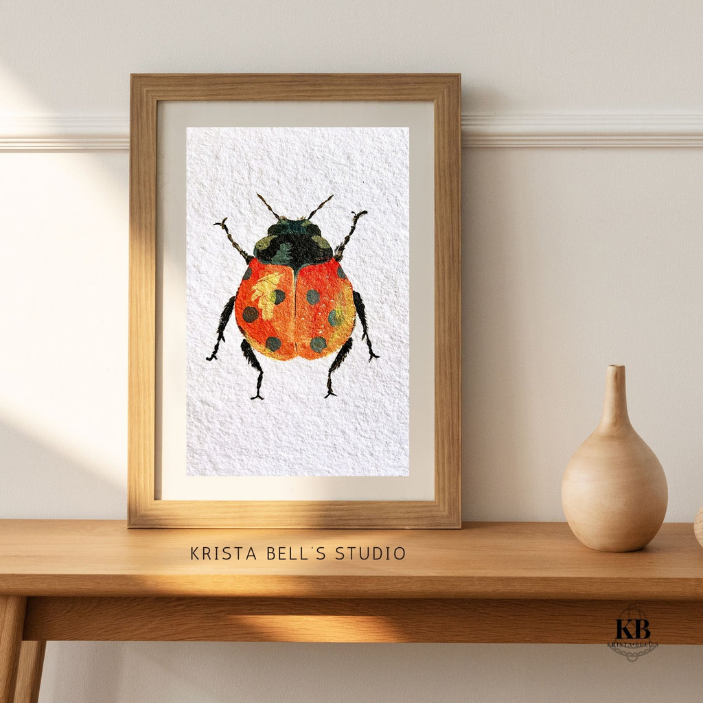 Framed artwork of a ladybug by Krista Bell's Studio on a wooden  shelf. A ladybug with reddish orange wings featuring black spots and a yellow highlight on a textured white background.