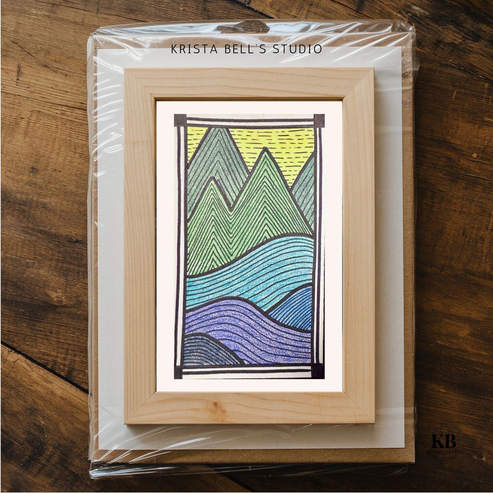 Framed artwork of mountains in green, yellow, and blue on a wooden surface with 'Krista Bell's Studio' branding.