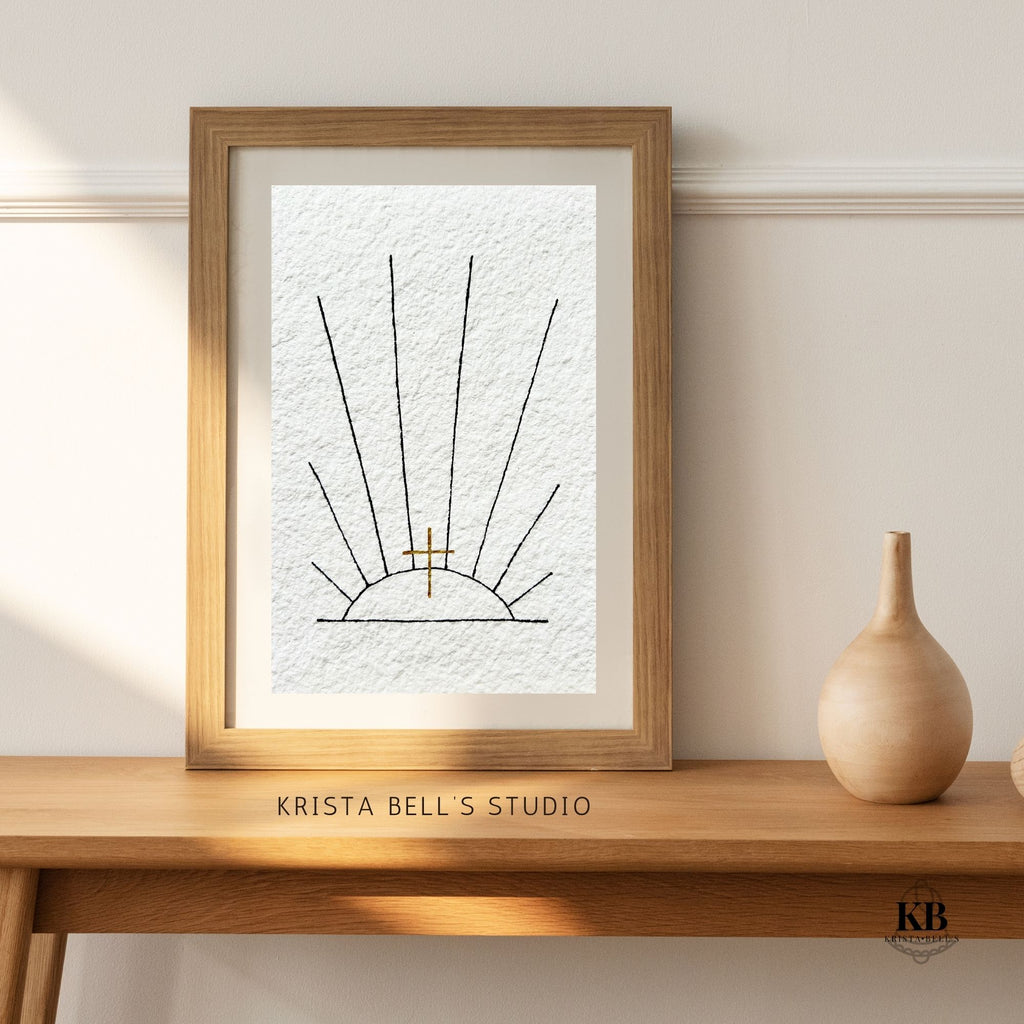 Framed artwork of black ink sunrays and a golden cross, on a wooden shelf with 'Krista Bell's Studio' branding.
