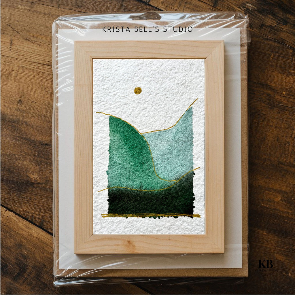 Framed artwork of simple modern watercolor landscape on a wooden surface, branded 'Krista Bell's Studio'.