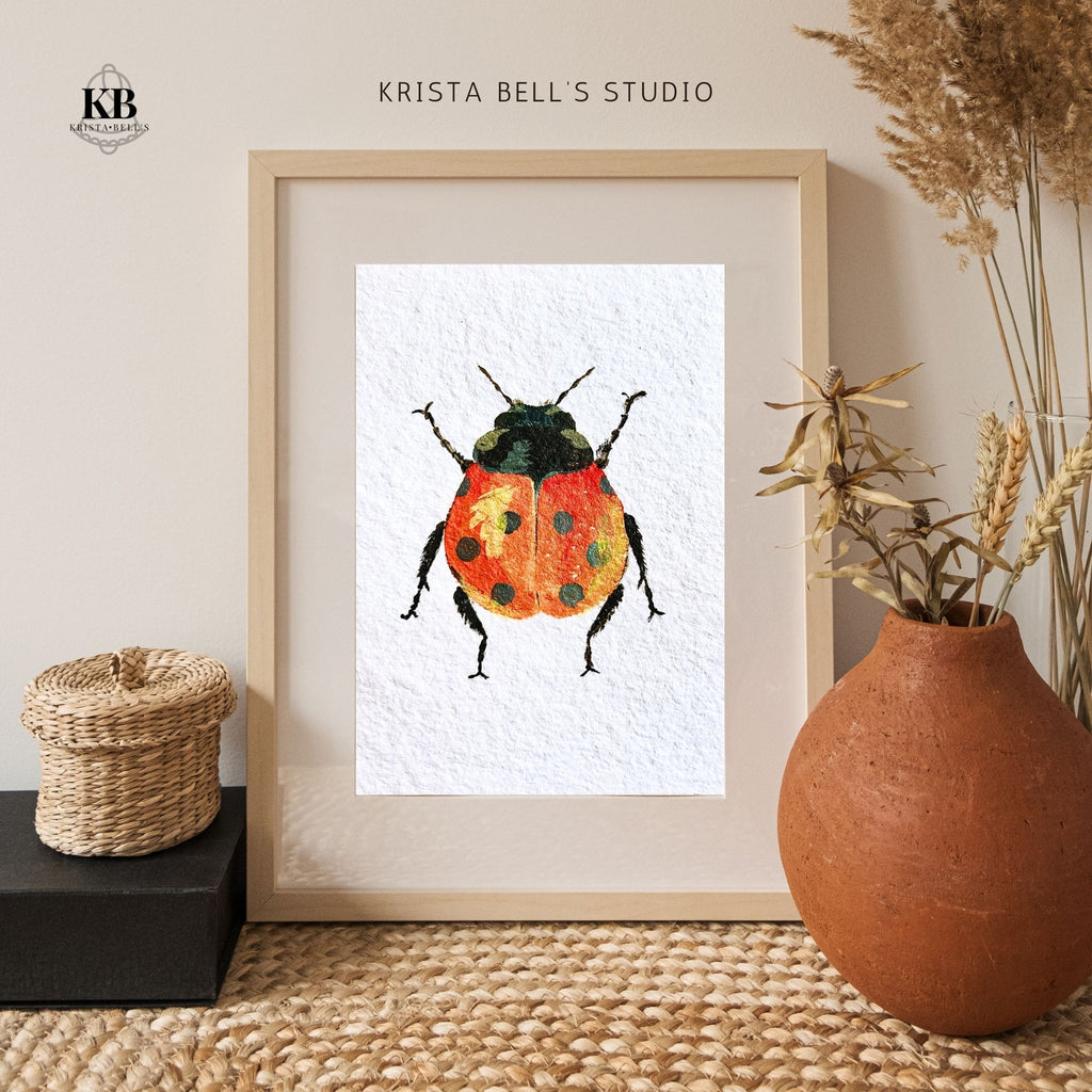 Framed artwork of a colorful beetle by Krista Bell's Studio, displayed on a neutral background with decorative elements. A ladybug with reddish orange wings featuring black spots and a yellow highlight on a textured white background.