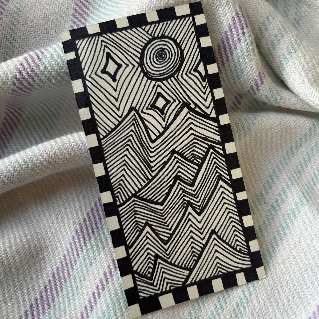 Black and white geometric patterned box bordering a geometric line landscape of jagged mountains and moon light, on a textured fabric background