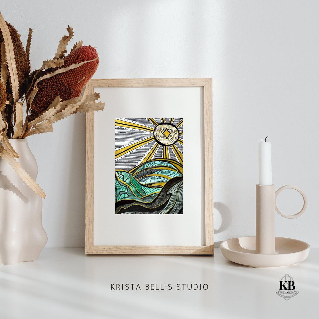 Framed artwork of a graphic landscape and golden rays on a white surface with decorative elements, featuring 'Krista Bell's Studio' branding.