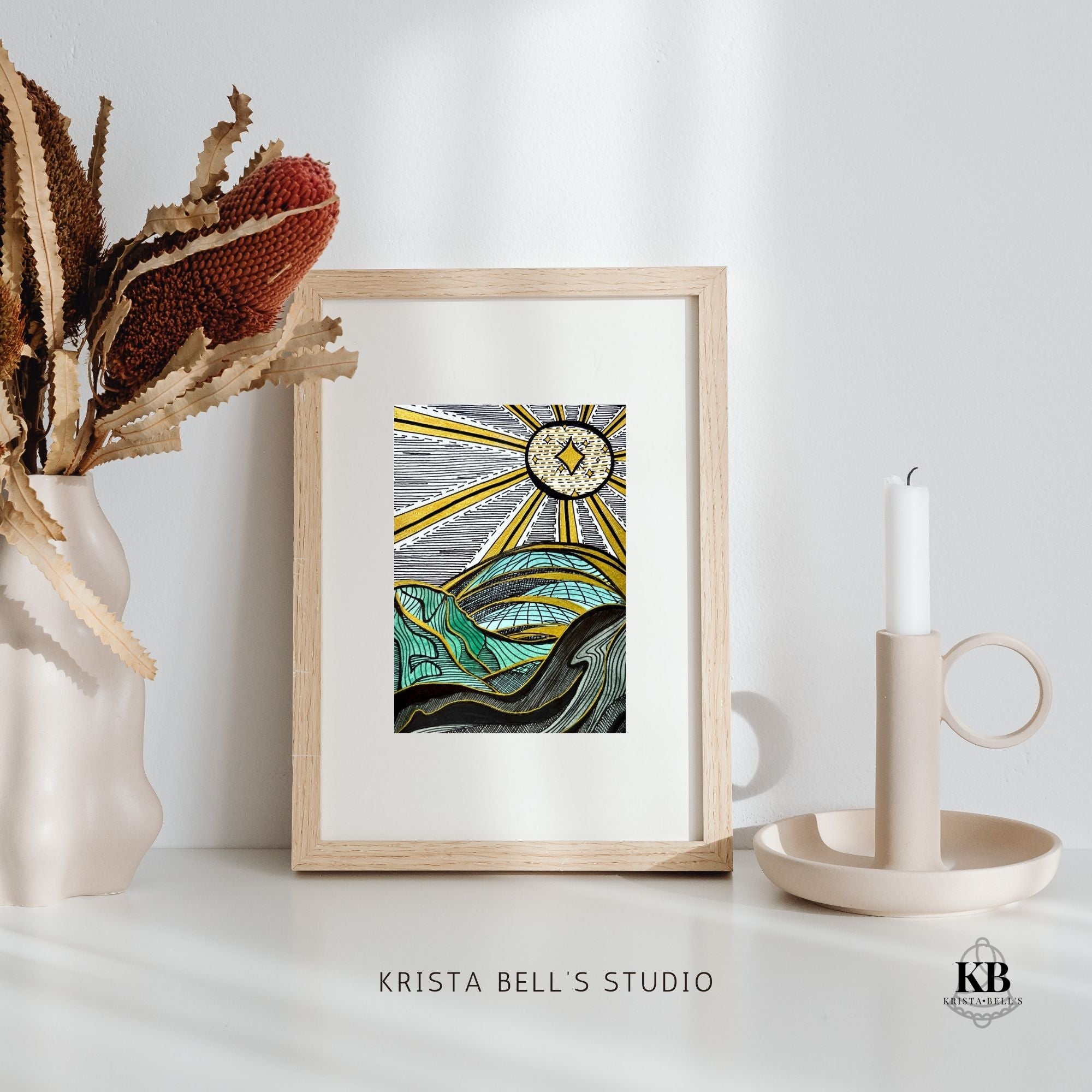 Framed artwork of a graphic landscape and golden rays on a white surface with decorative elements, featuring 'Krista Bell's Studio' branding.