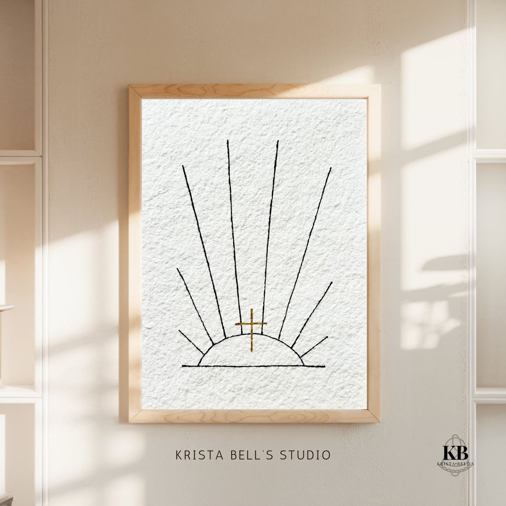 Framed artwork with black ink sunrays and a golden cross, on sun drenched wall, featuring 'Krista Bell's Studio' branding.