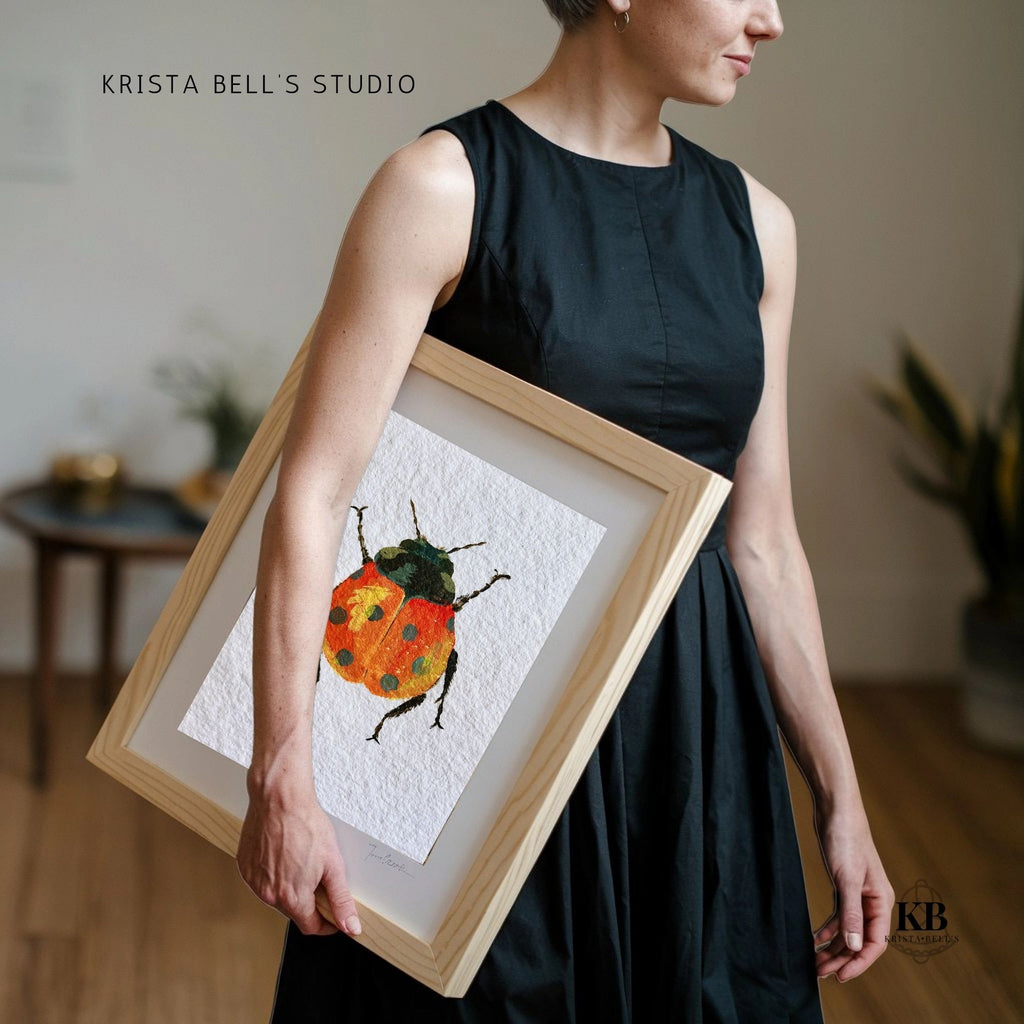 Person holding a framed artwork of a ladybug by Krista Bell's Studio. A ladybug with reddish orange wings featuring black spots and a yellow highlight on a textured white background.