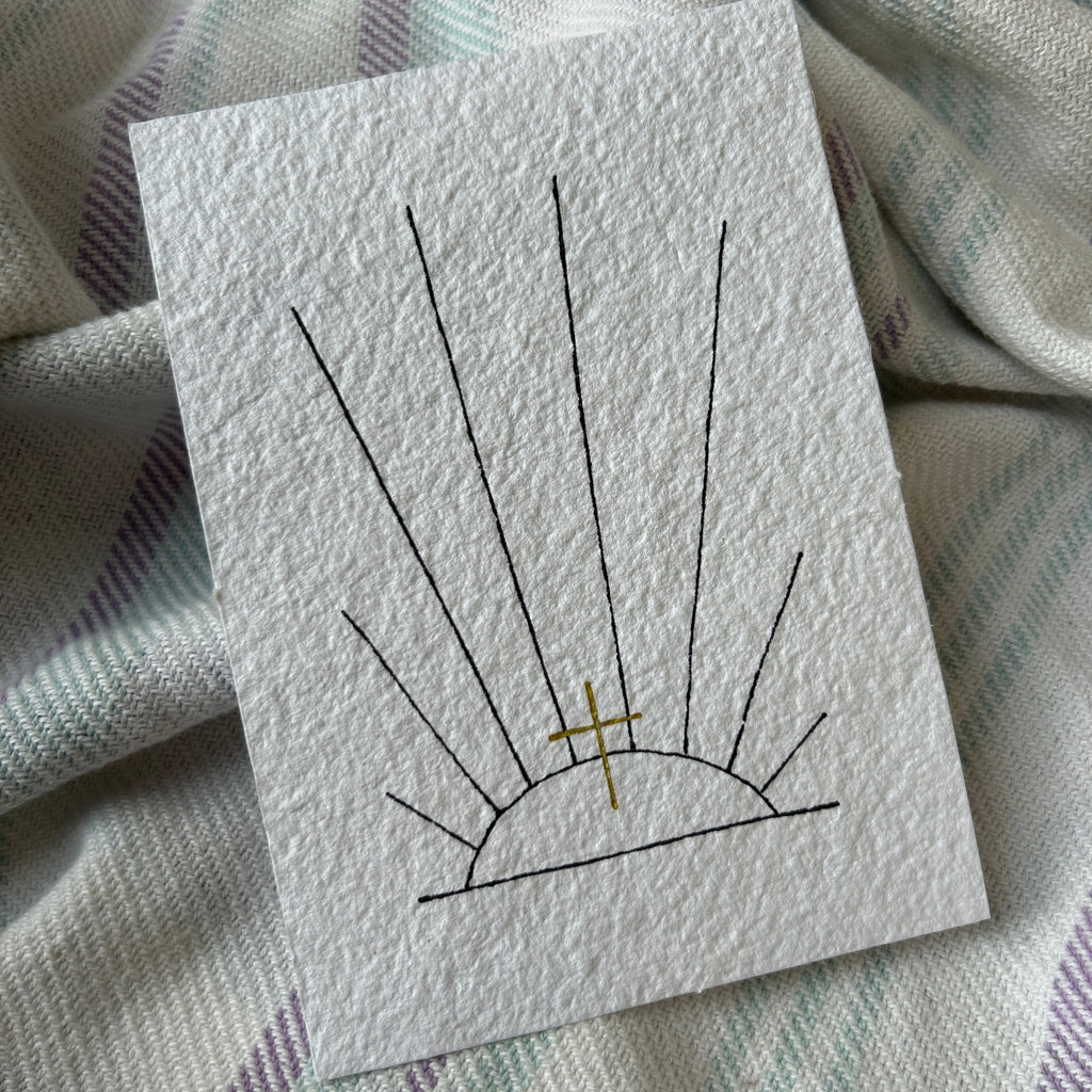 White card with black and gold design of black ink sunrays and a golden cross, on a textured fabric background