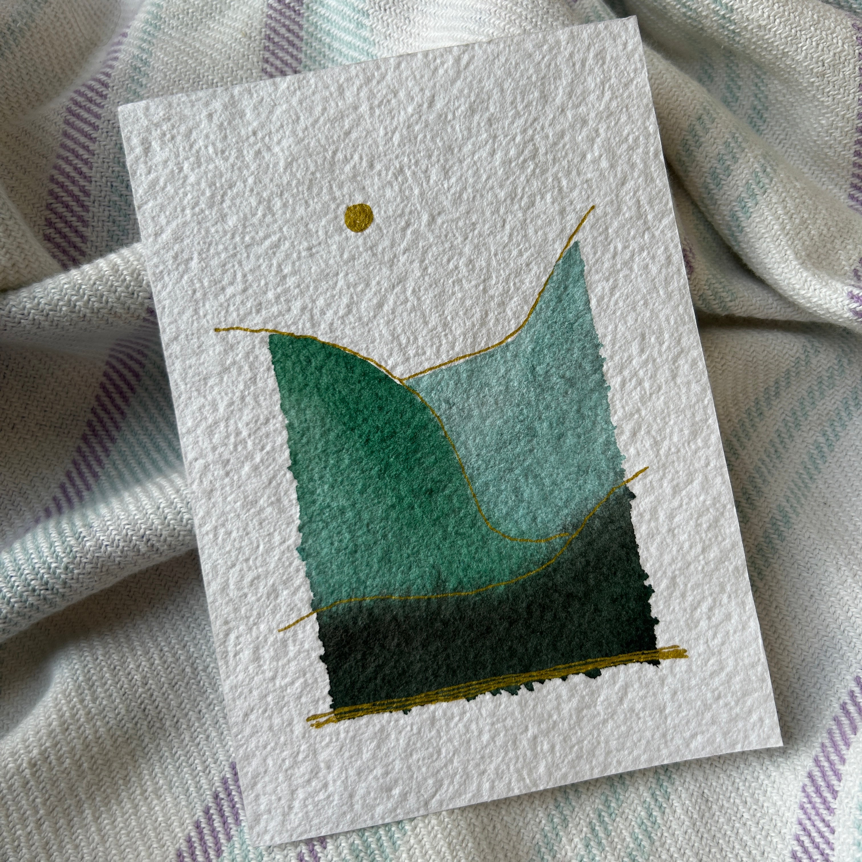 Handmade artwork with a simple modern watercolor landscape on textured paper, placed on a fabric background.