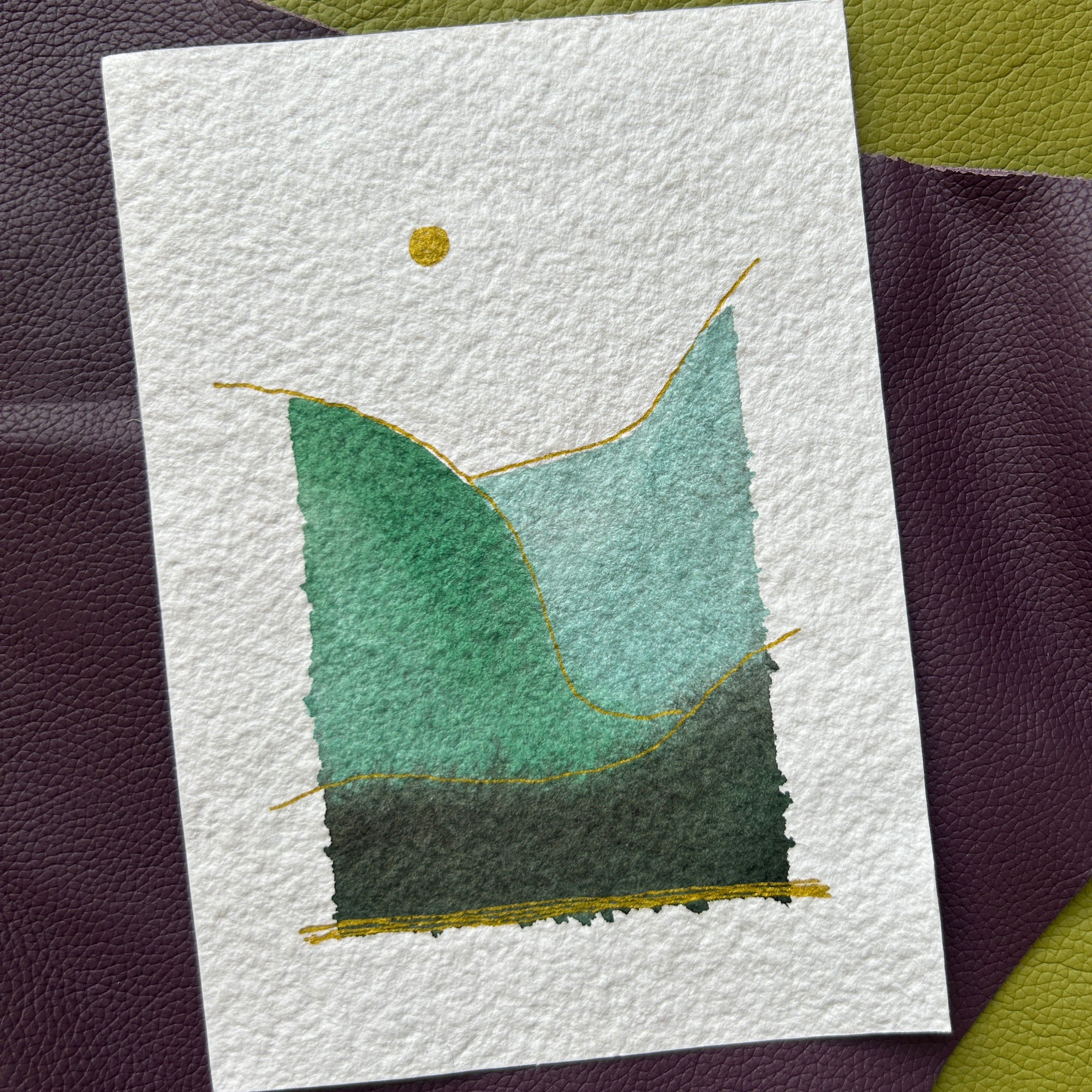 Handmade watercolor landscape card laying on a green purple background.