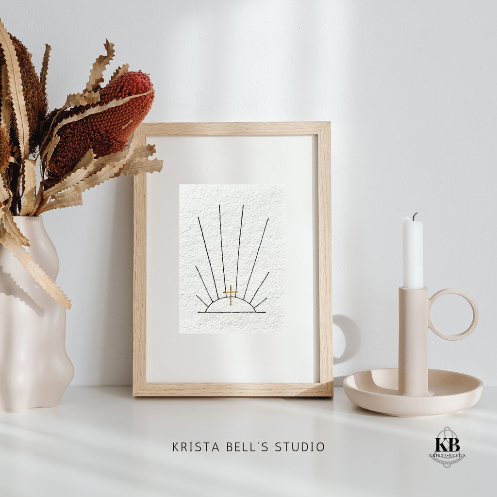 Framed artwork of sunrays and a golden cross on a white surface with decorative elements, featuring 'Krista Bell's Studio' branding.