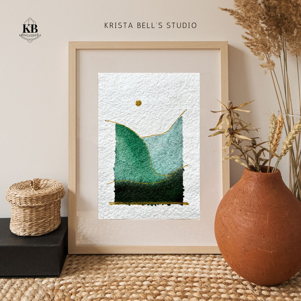 Framed simple modern watercolor landscape art piece with green and blue gradient, placed on a textured surface with decorative elements.
