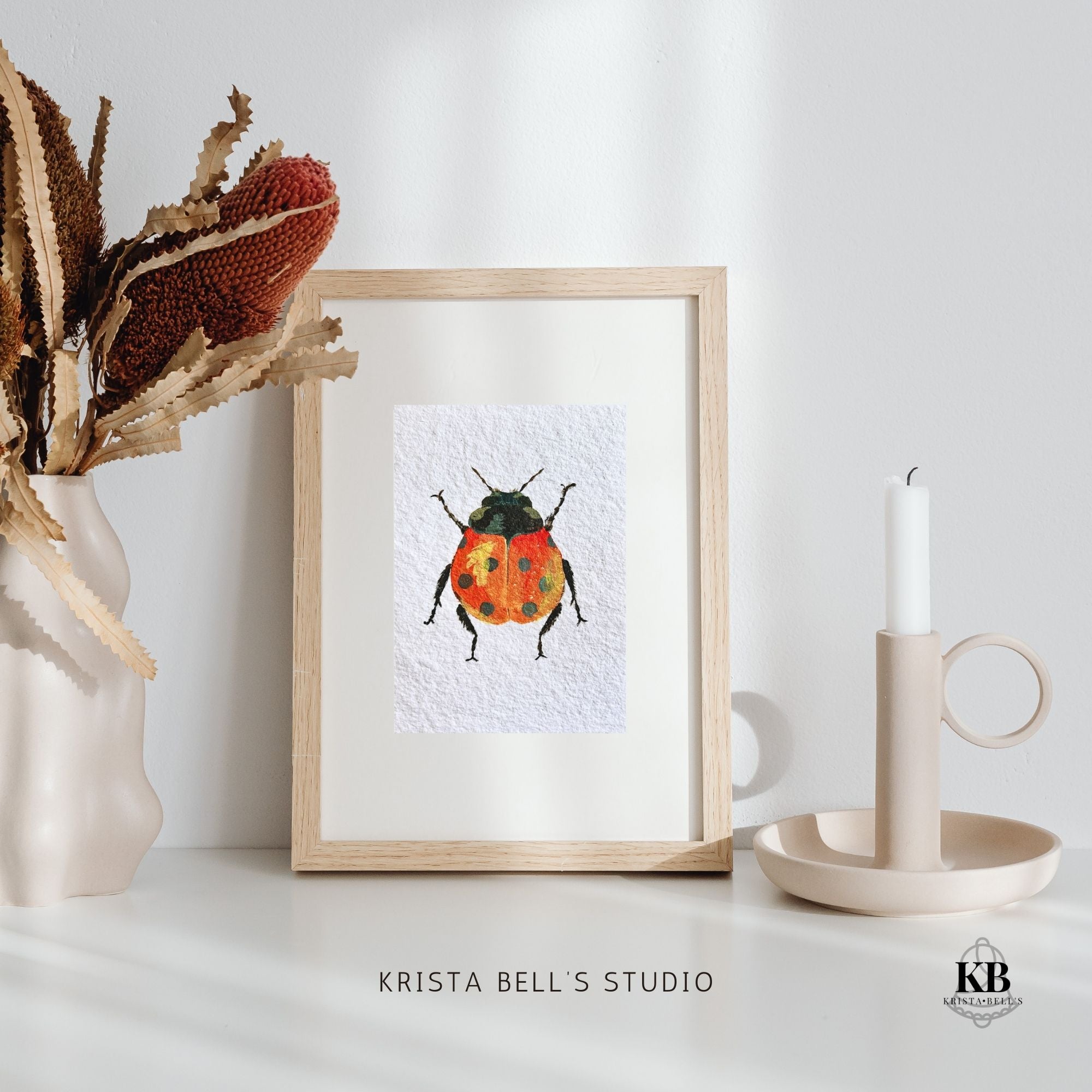 Framed artwork of a colorful beetle on a white surface with decorative items. A ladybug with reddish orange wings featuring black spots and a yellow highlight on a textured white background.