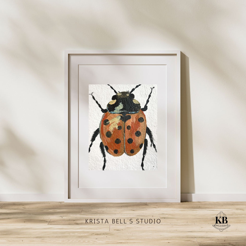 Framed artwork of a ladybug by Krista Bell's Studio on a wooden surface with a neutral background.