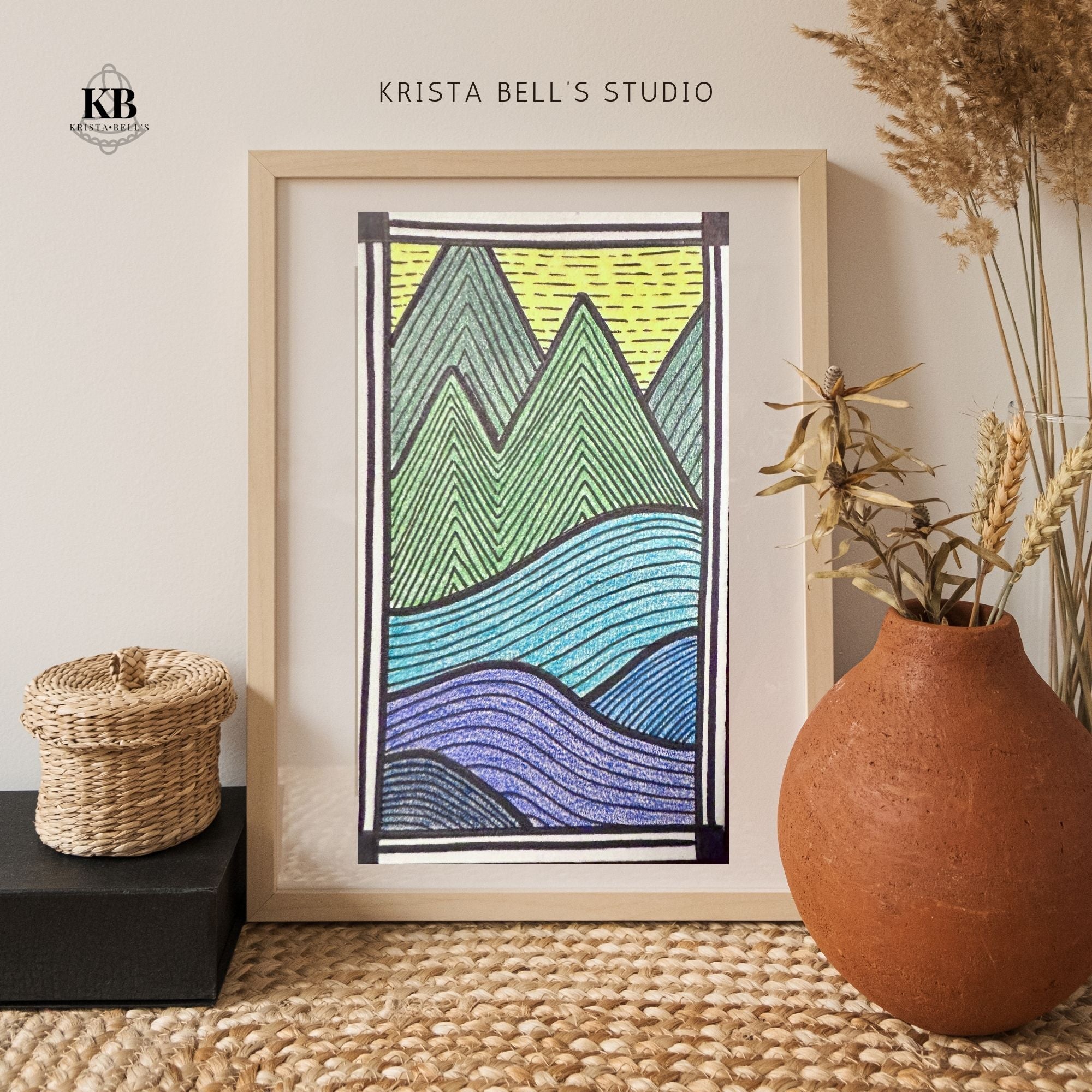 Framed abstract art piece with mountains, 'Krista Bell's Studio' logo, woven basket, and vase on a textured surface.