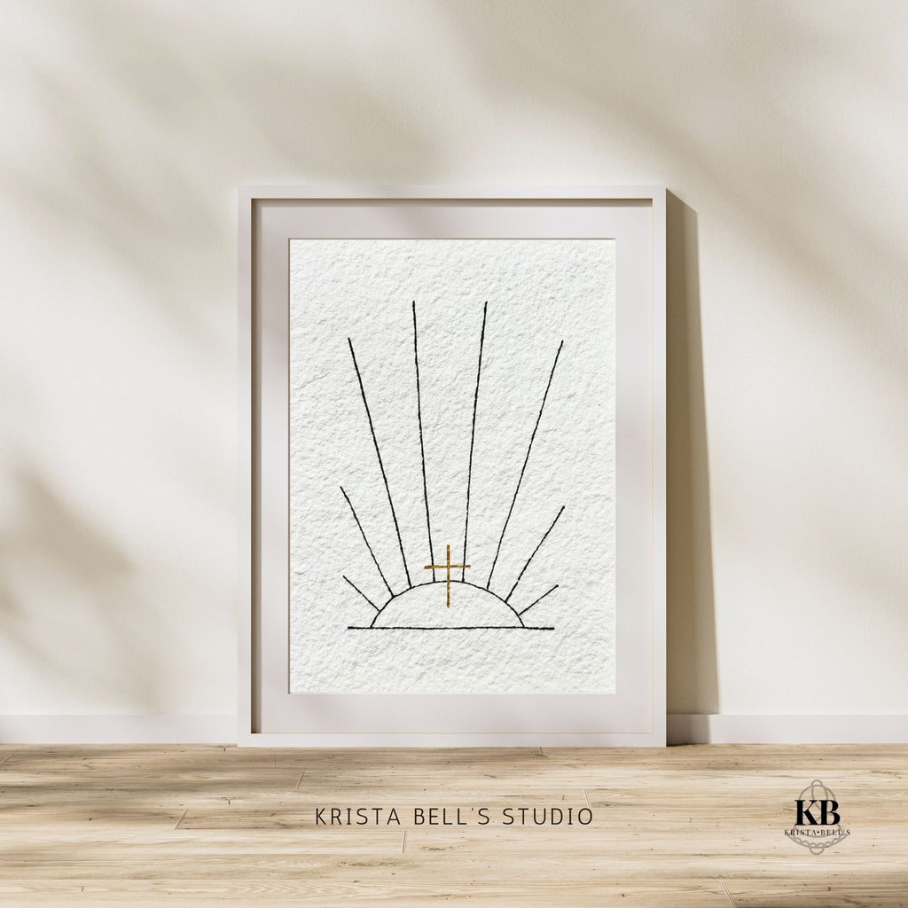 Framed original art of black ink sunrays and a golden cross on a wall with 'Krista Bell's Studio' branding.