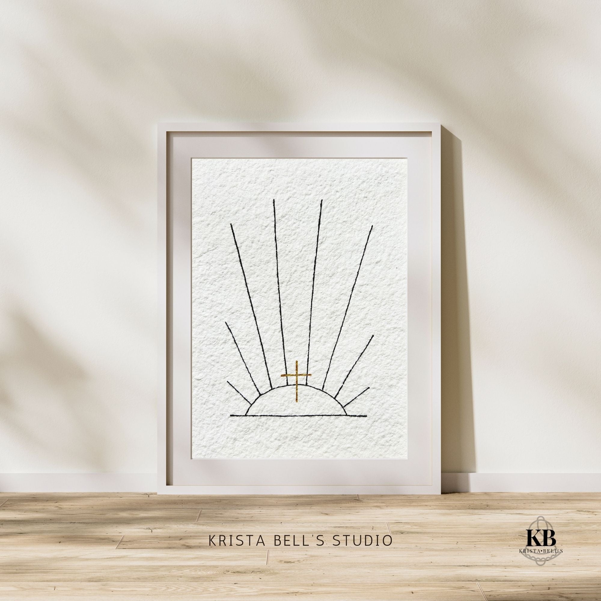 Framed original art of black ink sunrays and a golden cross on a wall with 'Krista Bell's Studio' branding.
