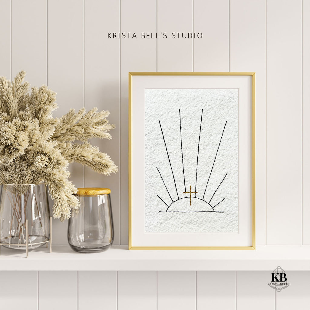 Framed artwork of black ink sunrays and a golden cross on a wall with decorative jars and dried plants.