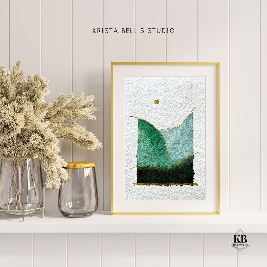 Framed simple modern watercolor landscape piece with a gold frame on a white wall, next to dried plants and a vase.
