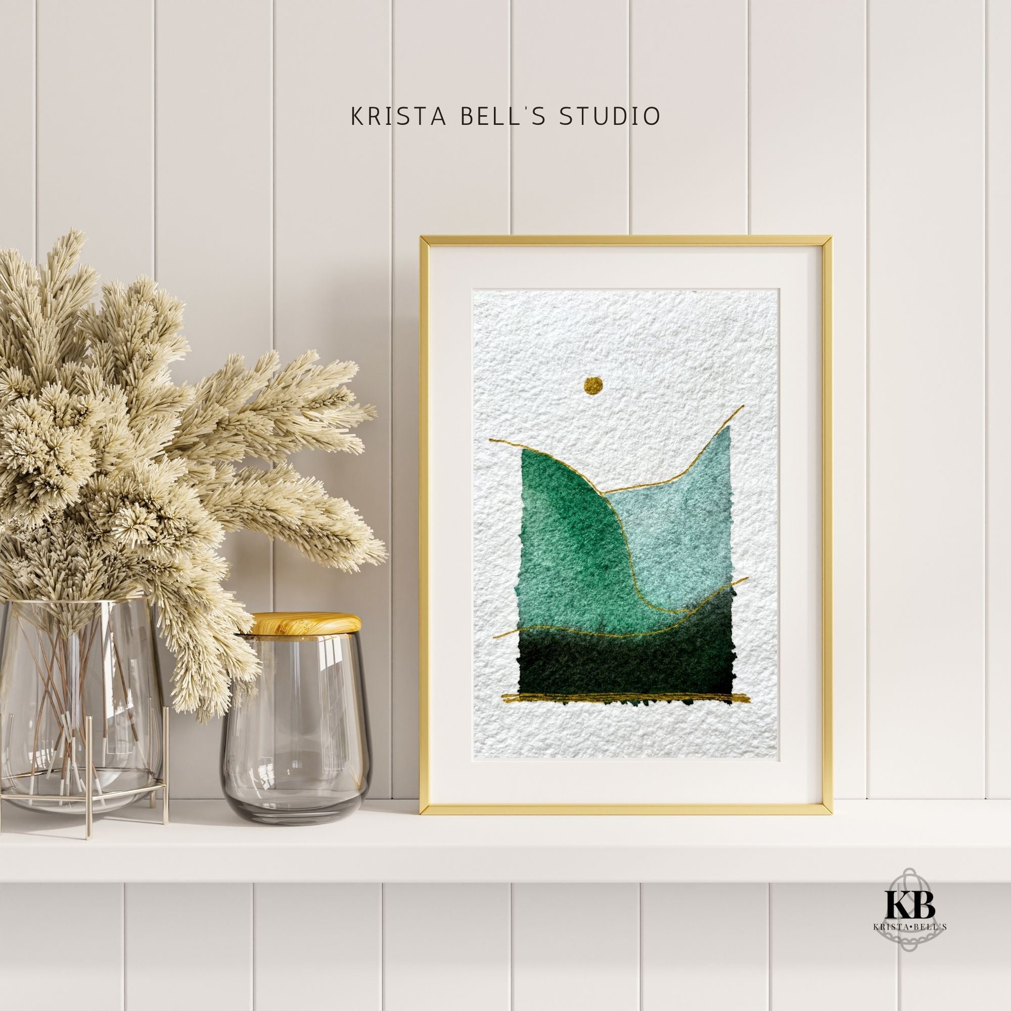 Framed simple modern watercolor landscape piece with a gold frame on a white wall, next to dried plants and a vase.