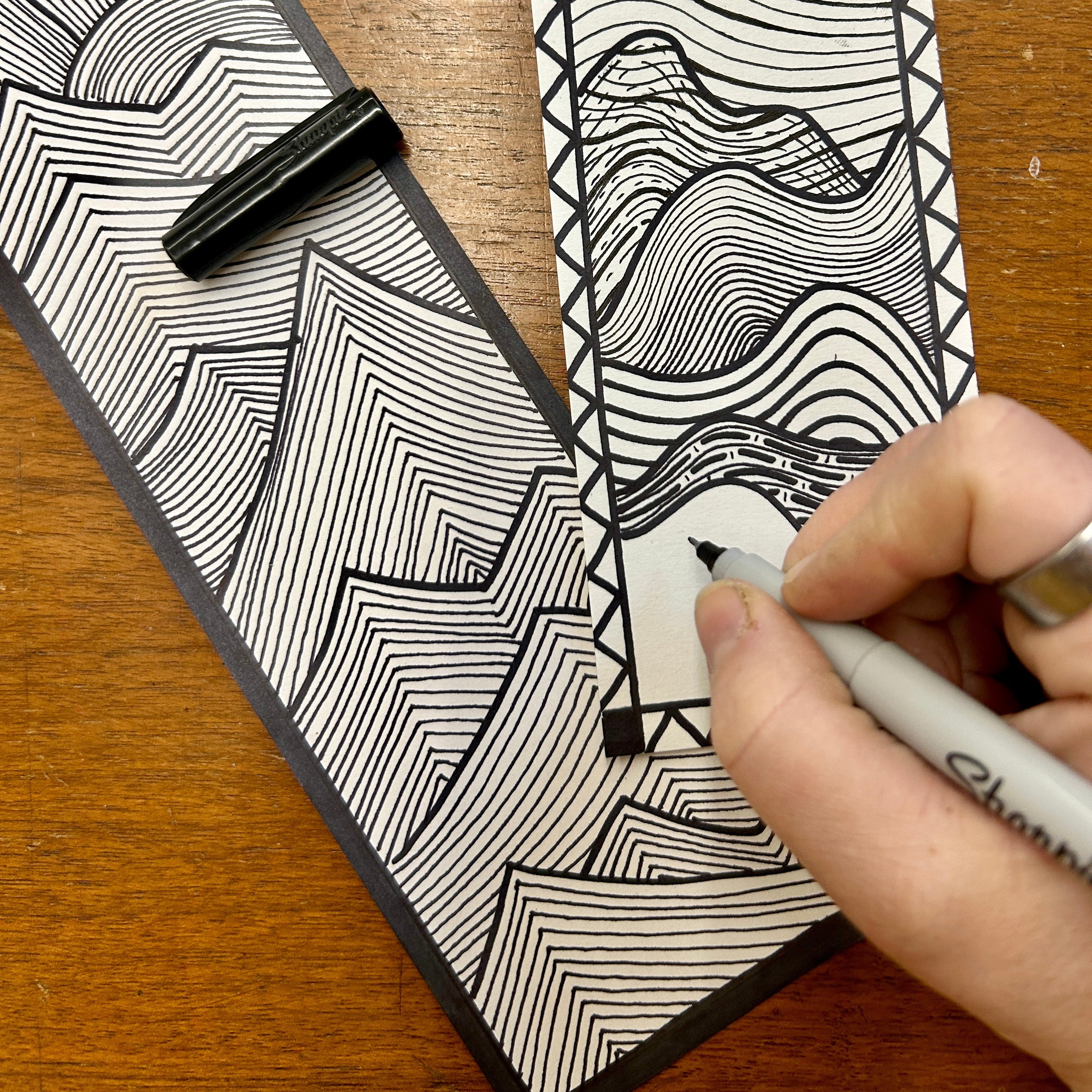 Person drawing a black and white line art design on a piece of paper with a pen.