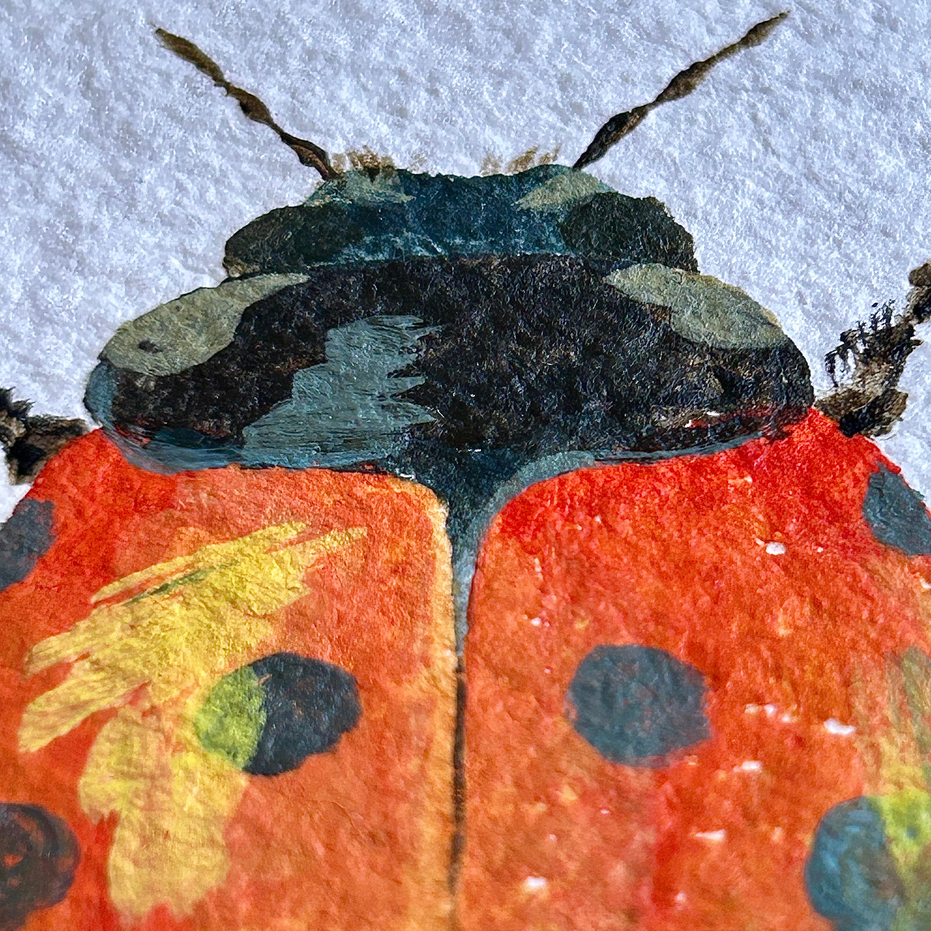 Close-up of a painted ladybug with red body, black spots, and yellowish brown antennae on a textured white background.