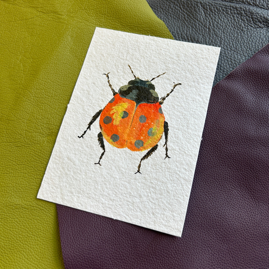 Painting of a ladybug on a white card with a textured surface. A ladybug with reddish orange wings featuring black spots and a yellow highlight on a textured white background.