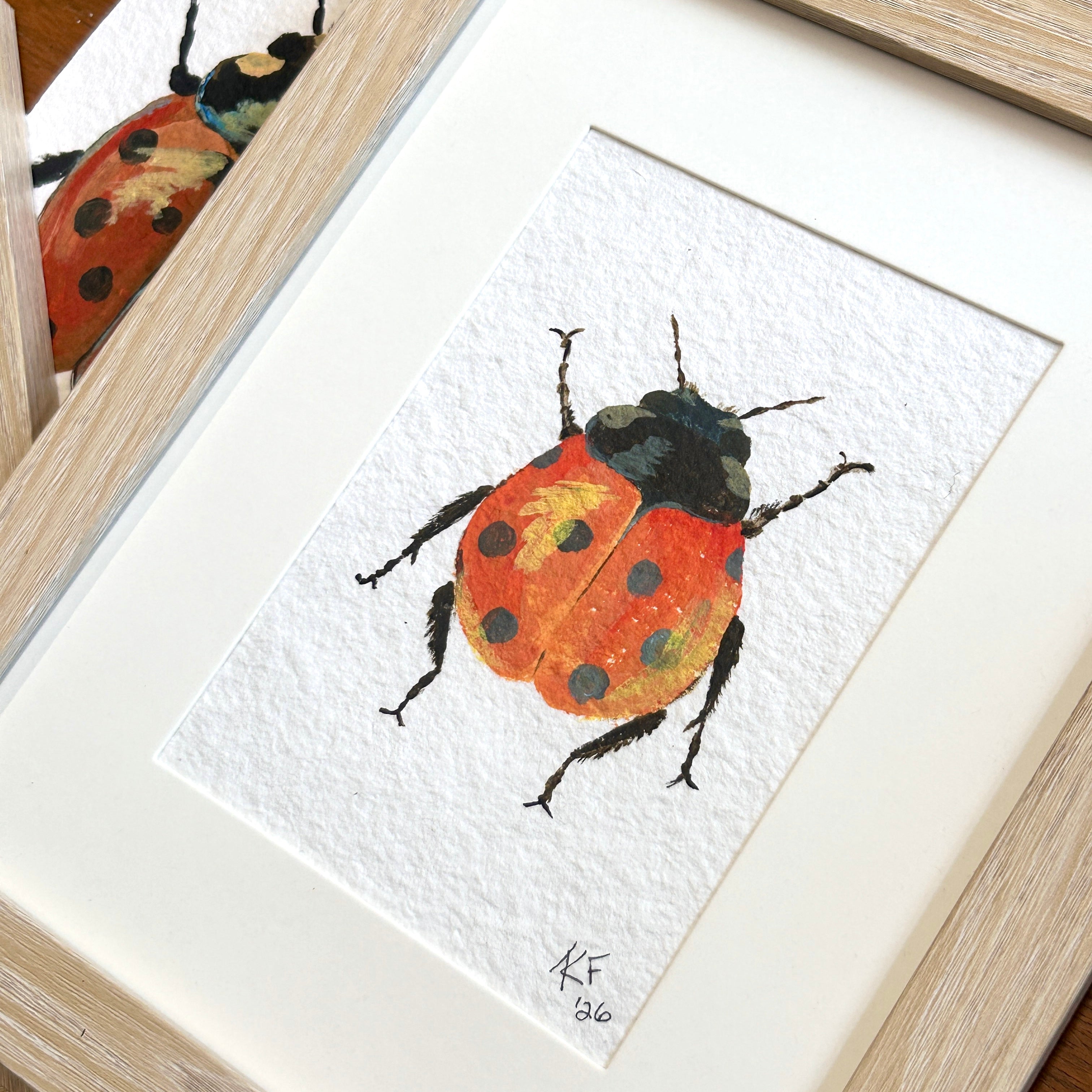 Framed watercolor painting of a ladybug on a textured white paper. A ladybug with reddish orange wings featuring black spots and a yellow highlight on a textured white background.