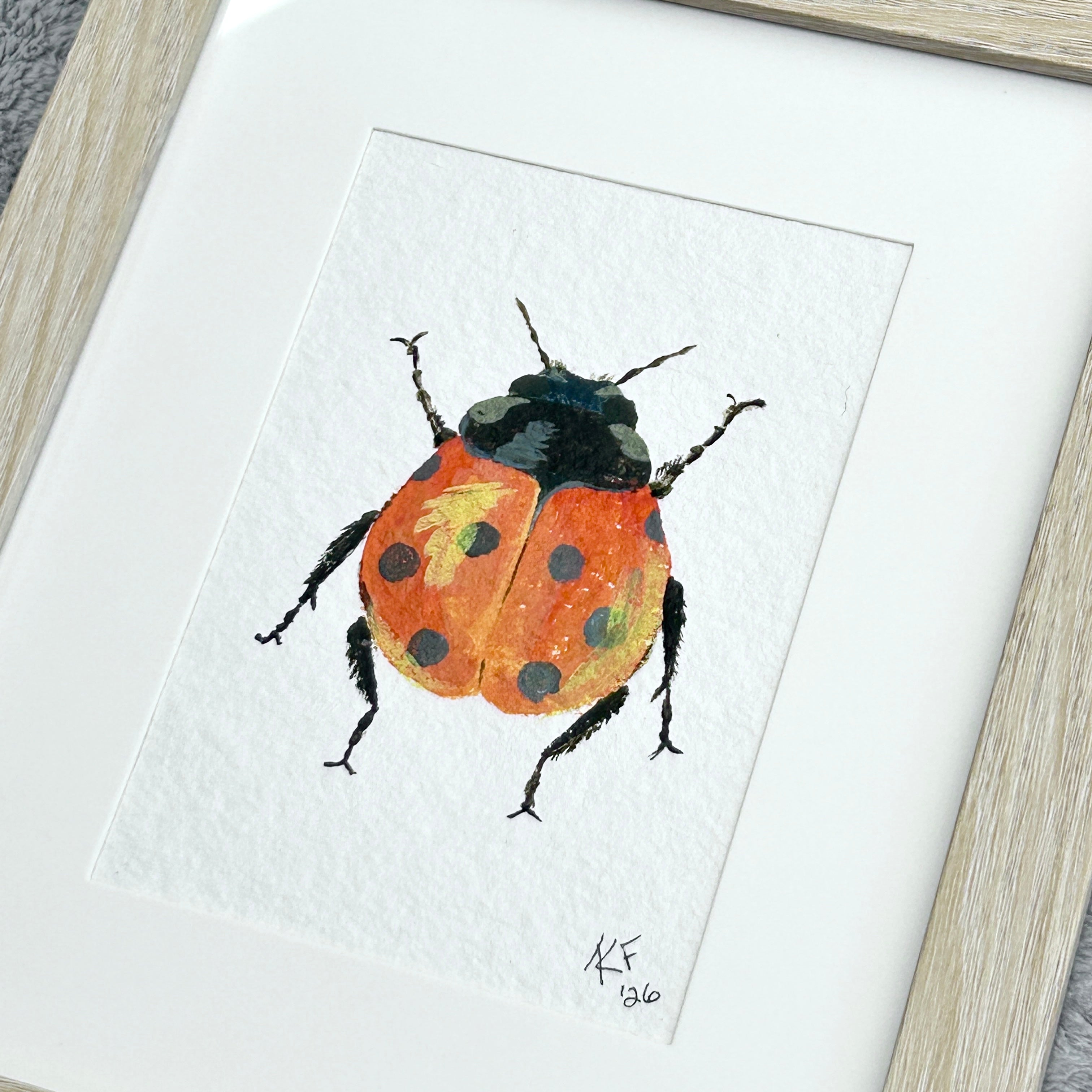 Framed acrylic painting of a ladybug with reddish orange wings featuring black spots and a yellow highlight on a textured white background.