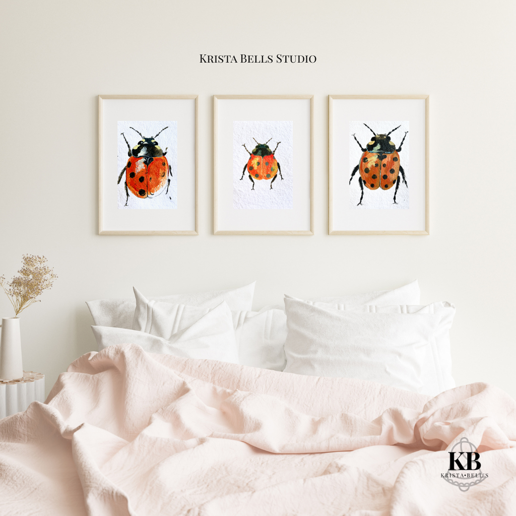 Three framed illustrations of ladybugs above a bed with pink bedding, featuring 'Krista Bells Studio' branding.